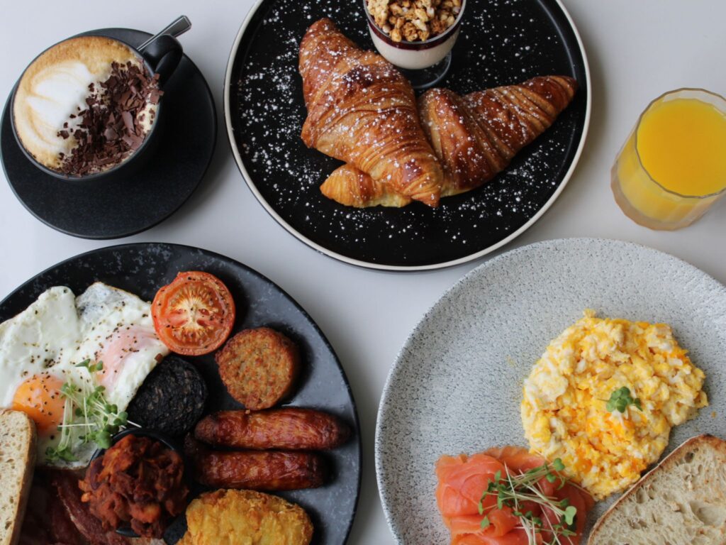 Aerial view of a breakfast spread: scrambled eggs with salmon, a full Irish breakfast, croissants with yogurt, cappuccino, and orange juice. Cozy and inviting.