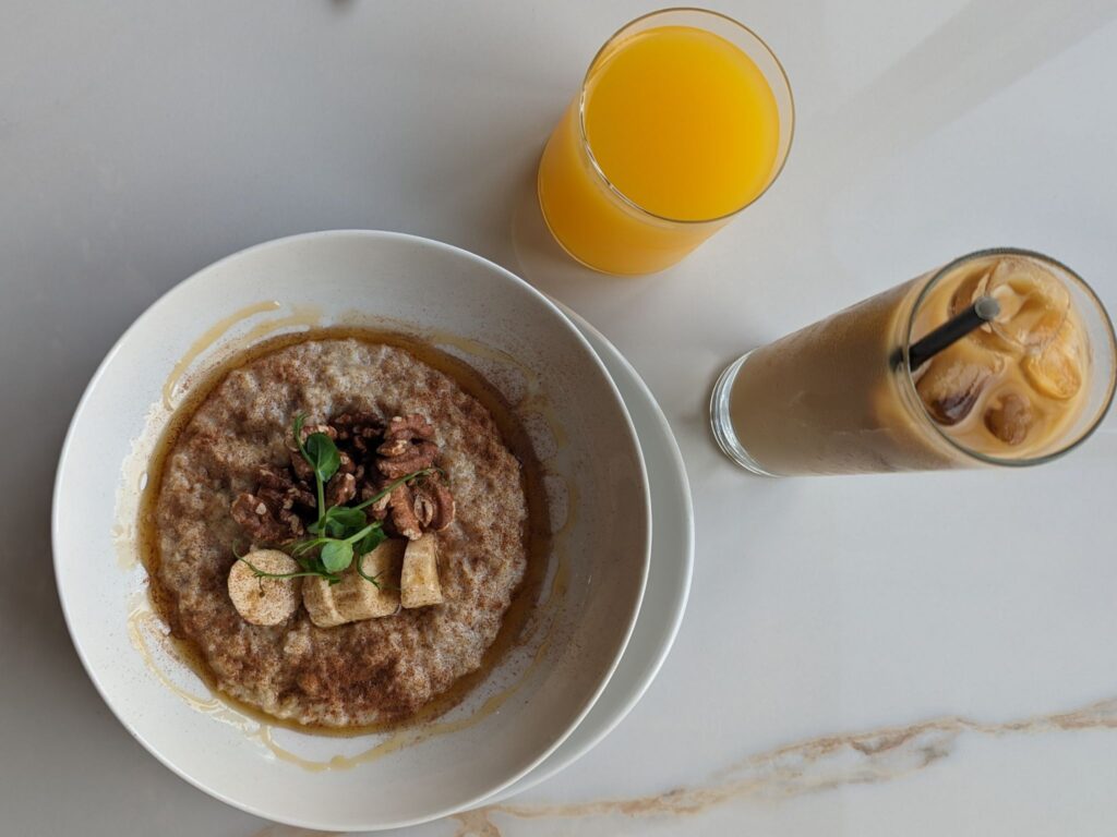Bowl of oatmeal topped with banana slices, pecans, and greens, served alongside a glass of orange juice and a glass of iced coffee with a straw on a light marble surface.