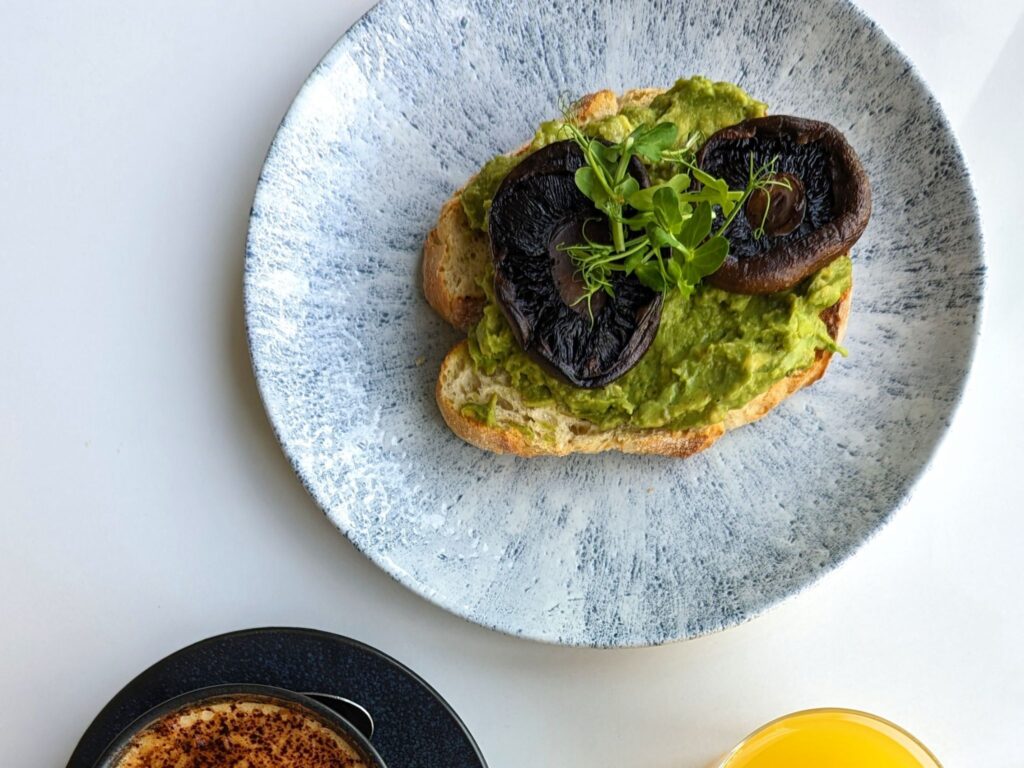 Avocado toast topped with mushrooms and greens on a textured plate. A cup of coffee and glass of orange juice are partially visible, evoking a fresh breakfast.