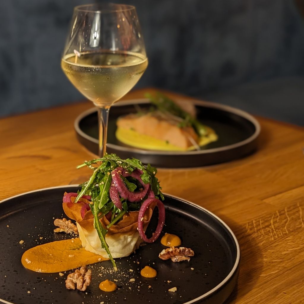 Close-up of a plated gourmet dish topped with arugula and pickled red onions beside a smear of orange sauce and scattered walnuts, with a glass of white wine and another plated dish blurred in the background.