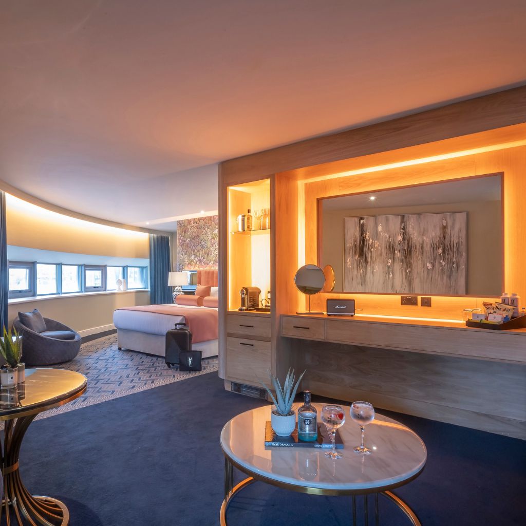 Spacious modern hotel suite with a curved wall of low windows, a pink bed in the background, and a seating area with a round table holding drinks and a small plant. A built-in wooden vanity with warm lighting and artwork is centered in the room.