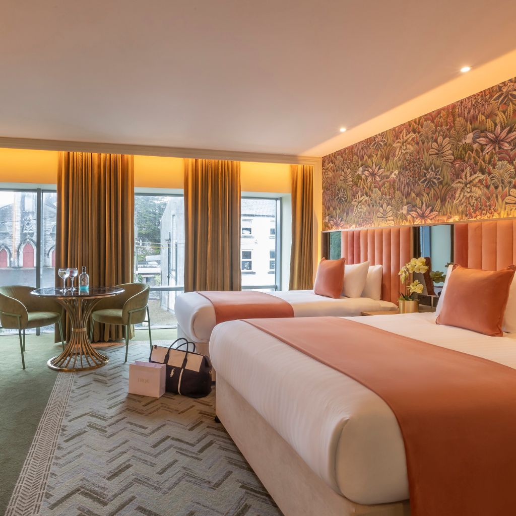 Bright modern hotel room with two large beds dressed in white linens and peach accents, set against a floral feature wall. Floor-to-ceiling windows with gold curtains line the wall, and a round glass table with green chairs sits by the window. Shopping bags and a travel bag are placed on the floor near the beds.