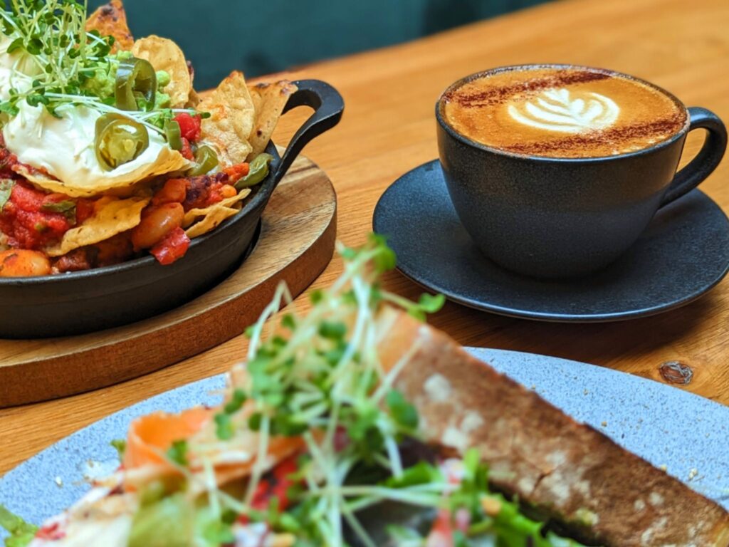 Tray of loaded nachos topped with sour cream, jalapeños, and greens next to a latte with foam art in a black cup; toasted sandwich in foreground.