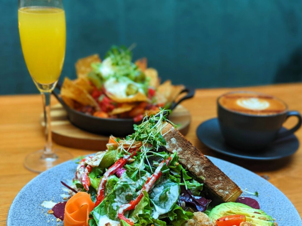 A fresh salad with greens, avocado, and toast on a plate, accompanied by a mimosa and cappuccino. A skillet with nachos sits in the background.