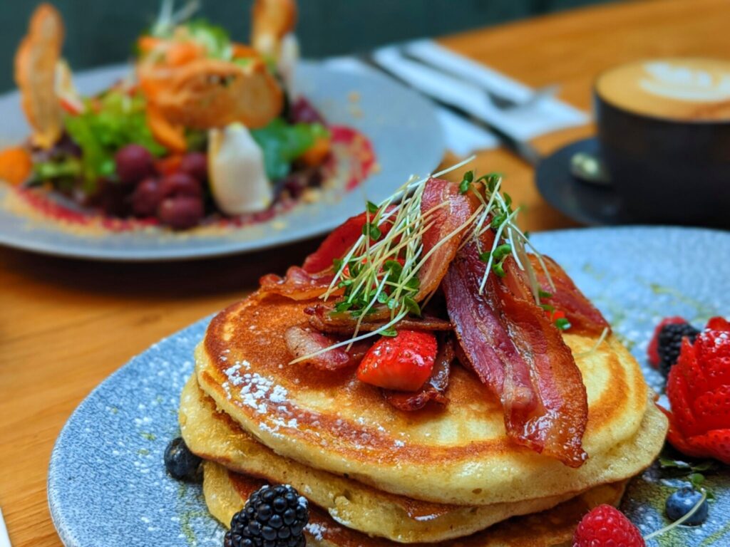 Stacks of fluffy pancakes topped with crispy bacon, strawberries, and microgreens on a plate. A colorful salad and cappuccino sit in the background. Cozy brunch scene.