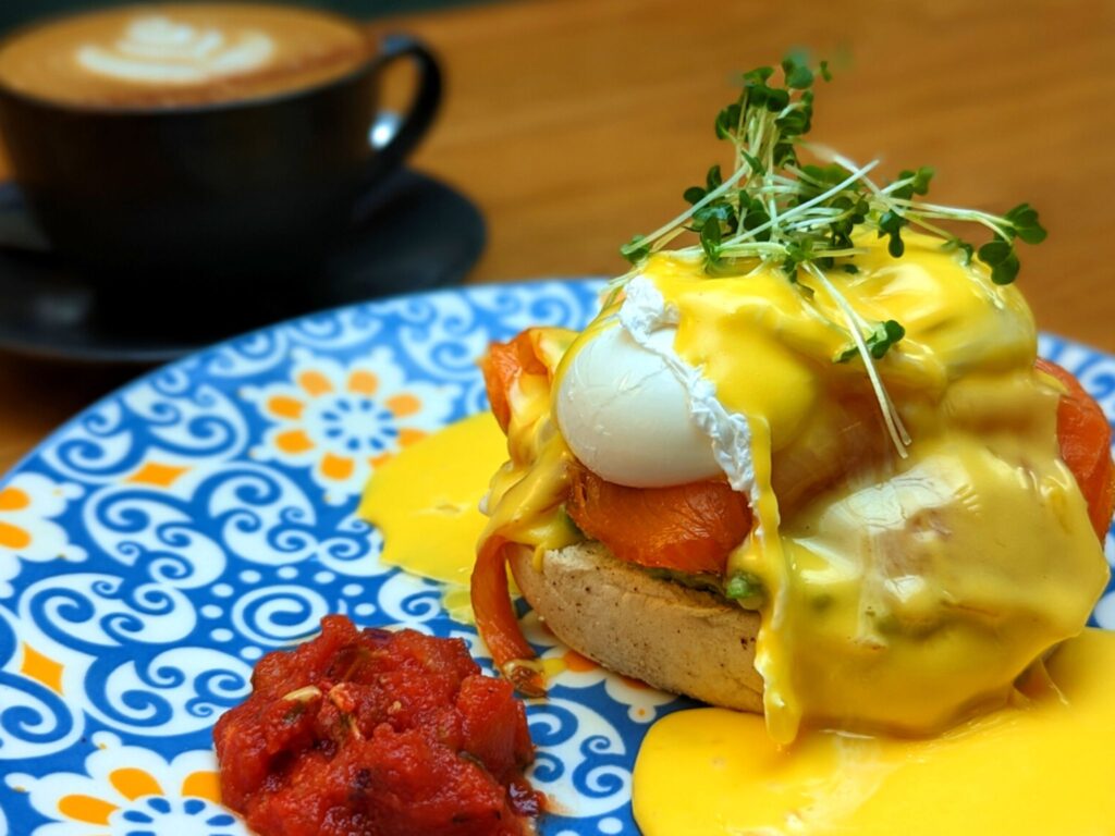 A vibrant plate with Eggs Benedict topped with creamy hollandaise, sprouts, and smoked salmon. Served with a side of red sauce. Cappuccino in the background.