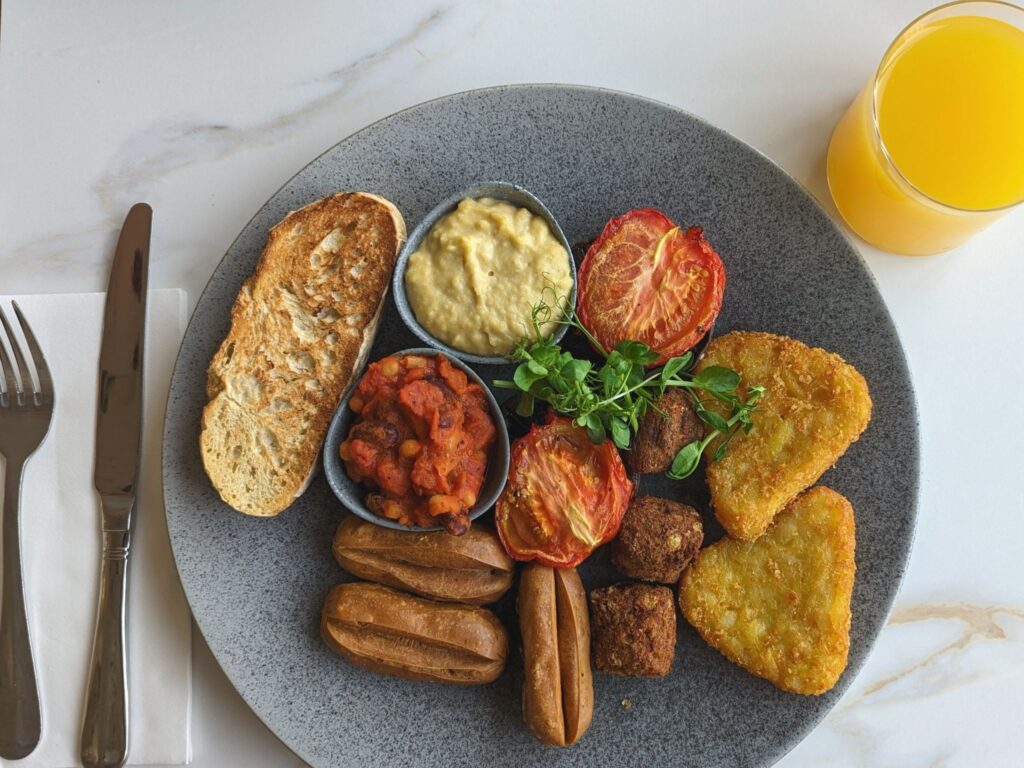 A gray plate with a full breakfast: toast, vegan sausages, baked beans, grilled tomatoes, hash browns, and hummus. A glass of orange juice is on the side.