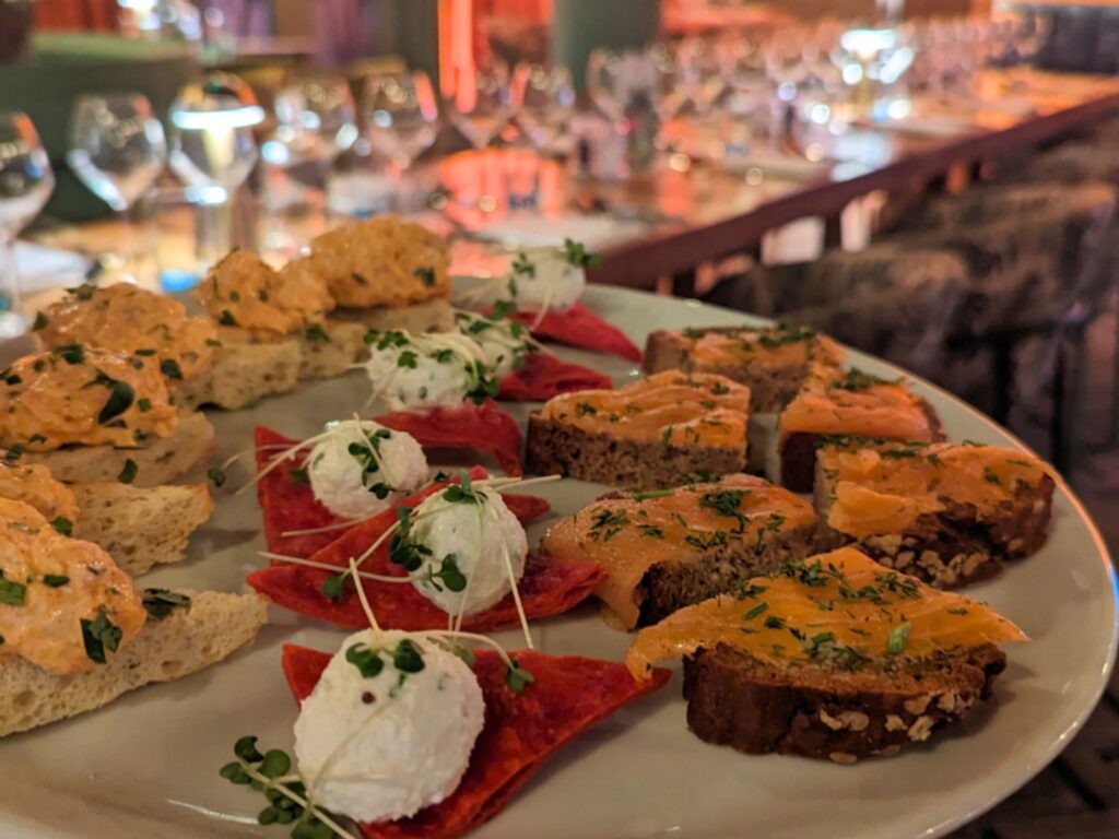 A close-up of an appetizer platter featuring assorted bite-sized canapés, including pieces of bread topped with a creamy orange spread, beetroot crisps with dollops of white cheese and microgreens, and slices of brown bread topped with smoked salmon and herbs. In the softly blurred background, an elegant table setting with wine glasses and cutlery is visible.