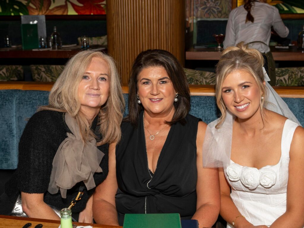 Three women sit closely together in a restaurant booth, smiling warmly at the camera. The woman on the right is dressed as a bride in a white gown with floral detailing, while the two women beside her wear elegant black outfits. The background shows a stylish, softly lit dining area.