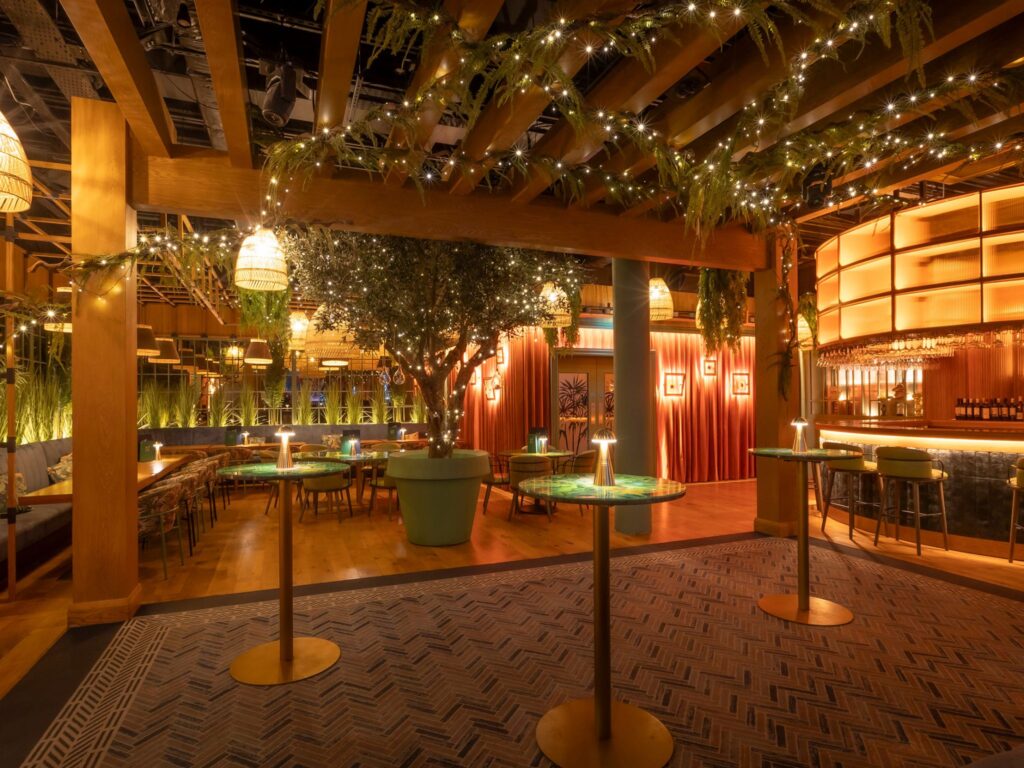 Warm, softly lit event space with wooden beams, hanging greenery, and string lights overhead. Several tall round cocktail tables with glowing lamps are spaced across the room. In the background, there are cushioned booths, lush plants, and a warmly illuminated curved bar area, creating an inviting and elegant atmosphere.