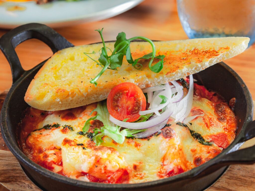 A sizzling skillet with melted cheese and tomato sauce is topped with a toasted bread slice, fresh tomato, onion rings, and greens. Warm and savory ambiance.