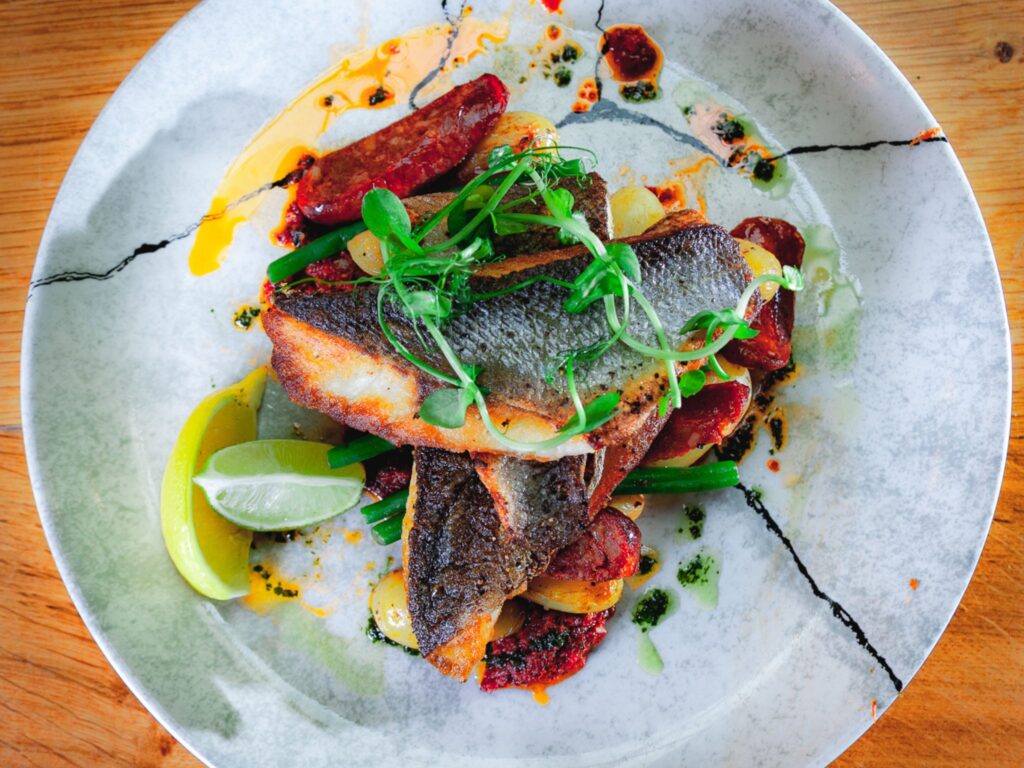 Grilled fish fillet on a cracked white plate with sautéed potatoes, vibrant greens, chorizo slices, microgreens, and lime wedges, exuding freshness.