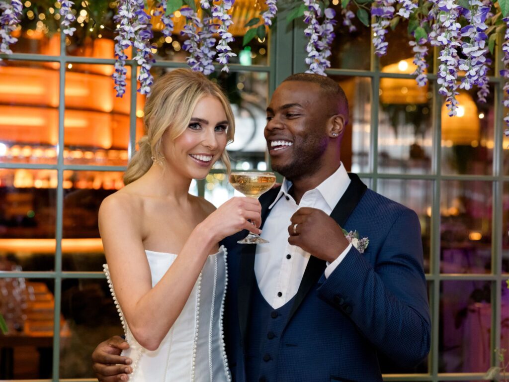 Bride and groom smiling and toasting with champagne glasses under hanging purple florals, standing in front of a window with warm ambient lighting in the background.