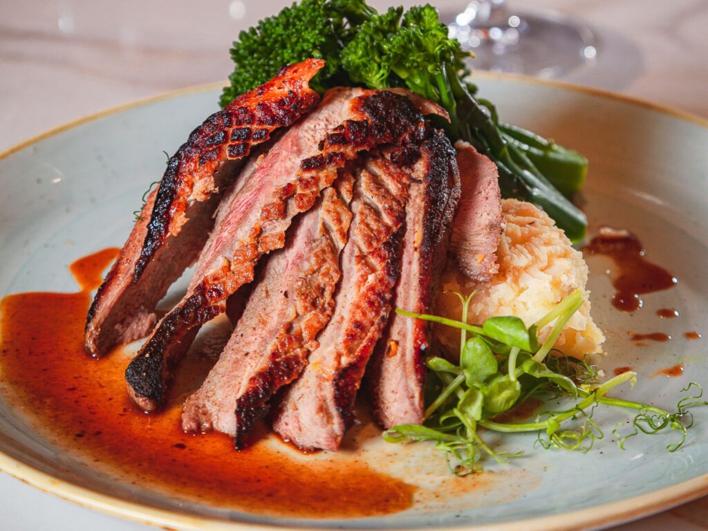 Succulent slices of grilled steak with a charred crust are served on creamy mashed potatoes, garnished with fresh greens and drizzled with rich sauce.
