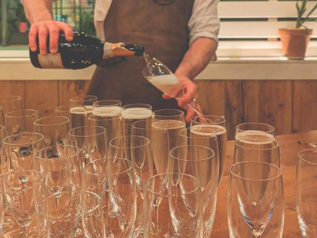 A server wearing an apron pours champagne from a bottle into a row of champagne flutes arranged on a wooden table.