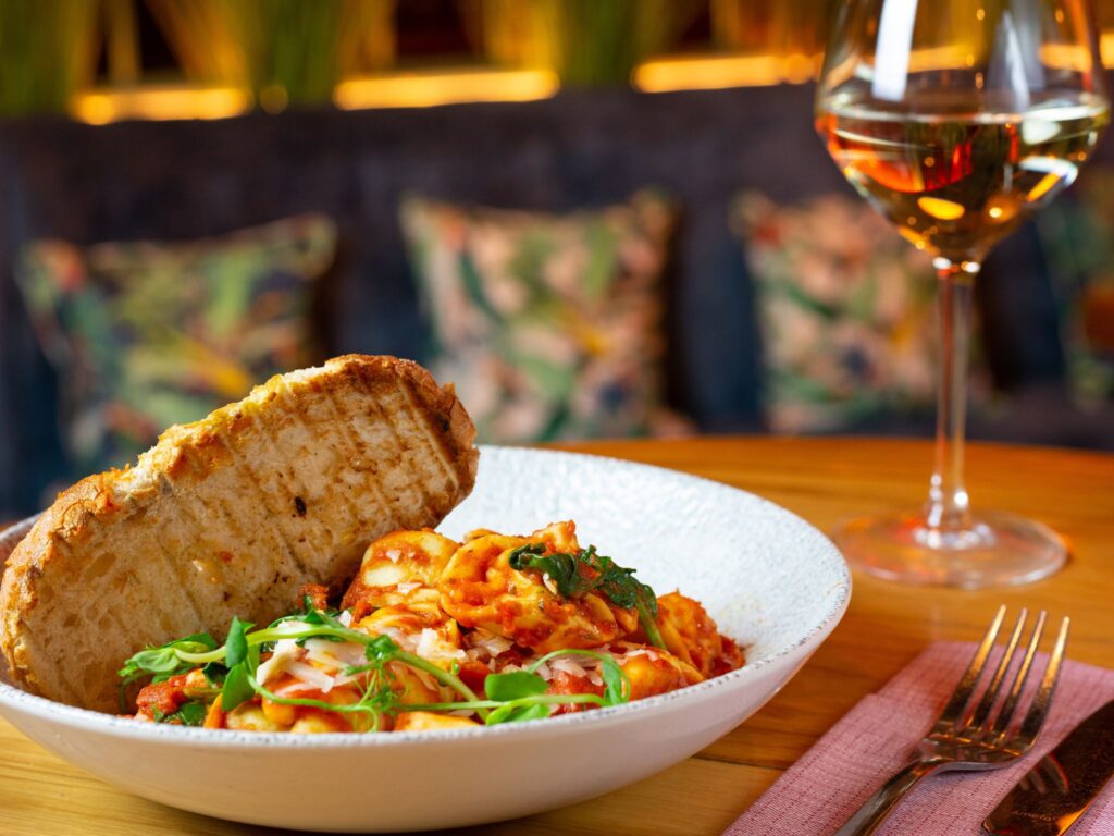 A bowl of pasta with tomato sauce, topped with greens and cheese, next to grilled bread. A glass of white wine sits nearby on a wooden table.