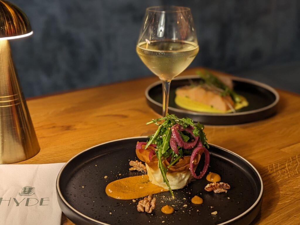 Elegant dining setup with a sleek black plate featuring a gourmet dish topped with arugula and pickled onions. A glass of white wine and a modern lamp enhance the ambiance.