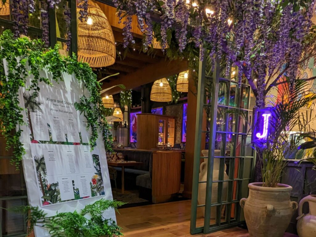 A wedding seating chart decorated with lush green foliage stands at the entrance of a warmly lit venue with hanging wicker lamps, purple wisteria, and large potted plants. A neon purple letter ‘J’ glows on the right side near a tall plant.