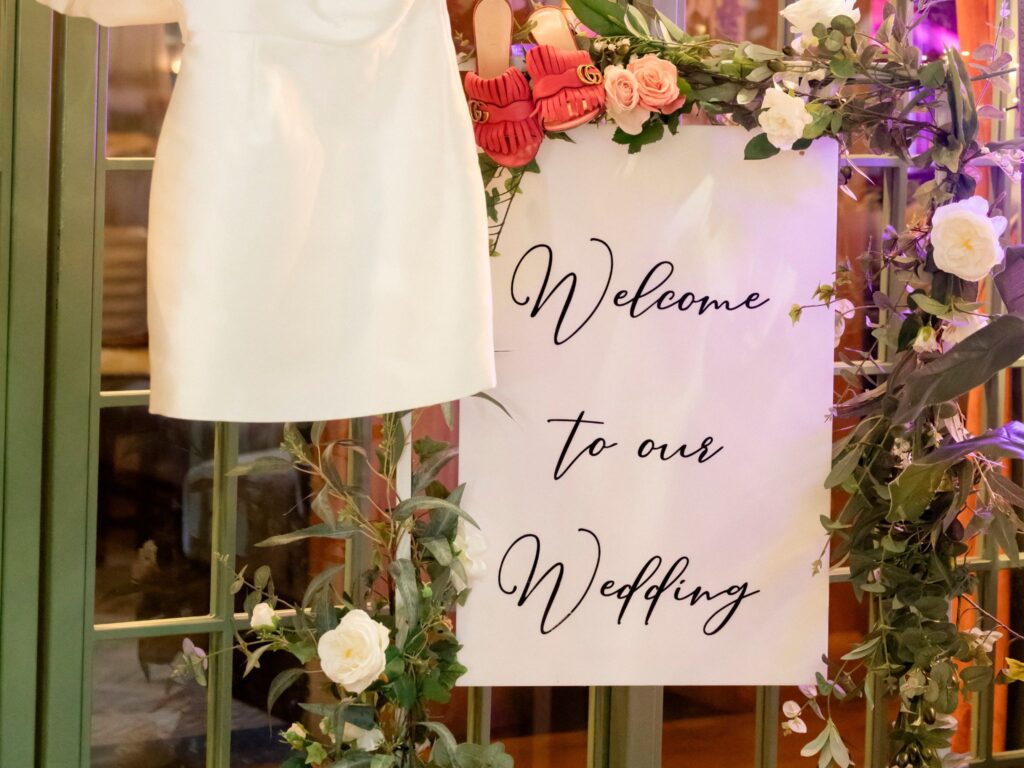 Decorative wedding sign framed with pink and white flowers and greenery, reading ‘Welcome to our Wedding,’ hanging beside a white dress and floral arrangements.