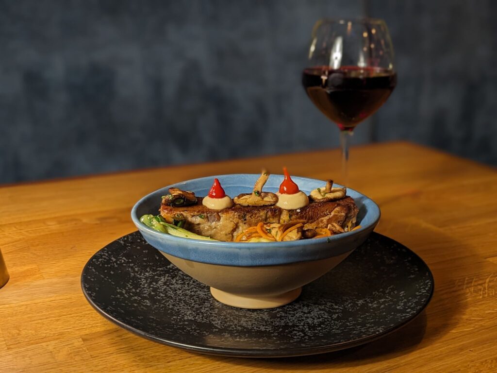 A gourmet bowl of noodles topped with mushrooms, sauces, and garnishes is placed on a wooden table. A glass of red wine is in the background.