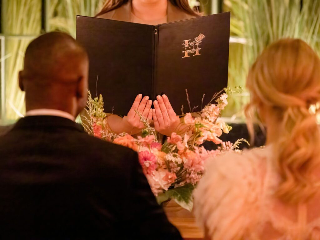A wedding officiant stands behind a floral arrangement, holding an open black folder with the Hyde logo, while a couple sits before her during the ceremony.