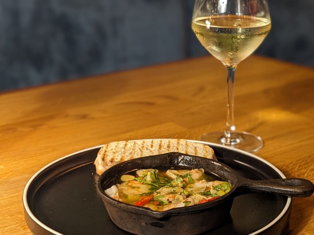 A black skillet with shrimp in a herbed sauce, accompanied by toasted bread, sits on a wooden table. A glass of white wine is nearby, creating a cozy dining scene.