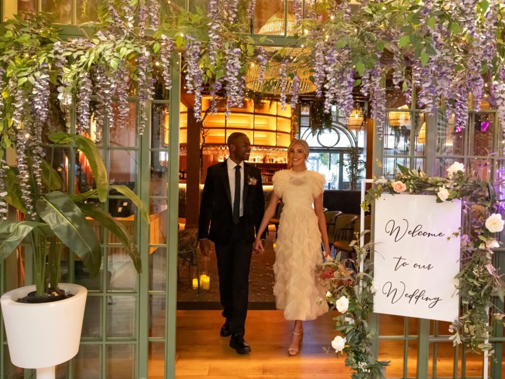 A bride and groom walk hand in hand through a floral-decorated entrance with hanging wisteria and greenery. A sign beside them reads “Welcome to our Wedding,” and the warmly lit venue interior is visible behind them.