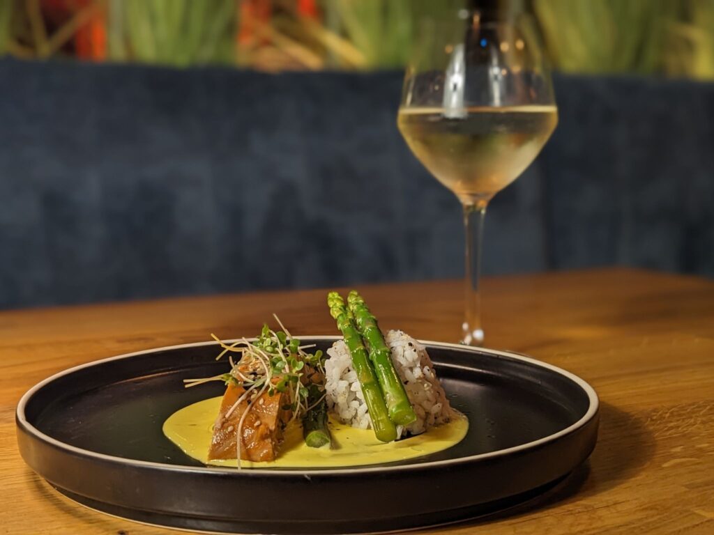 A stylish plate of gourmet food featuring asparagus spears on rice and glazed meat next to a creamy sauce, paired with a glass of white wine. Cozy ambiance.