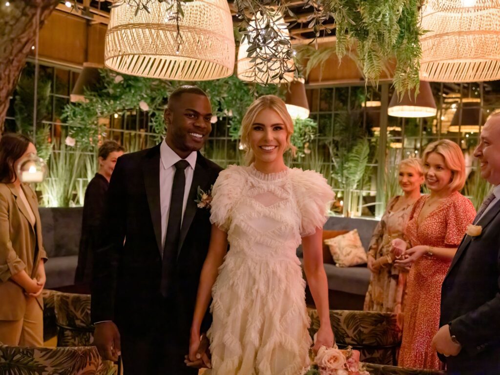 A newlywed couple walks hand in hand indoors, smiling warmly as guests stand nearby in a lush, plant-filled, softly lit venue.
