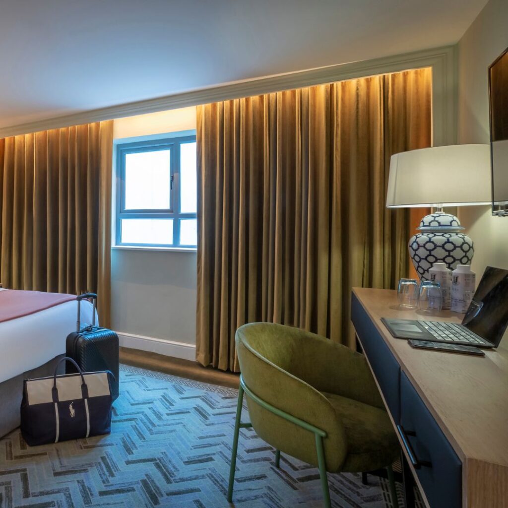 A stylish hotel room featuring a modern desk with a laptop, lamp, and water glasses. A green velvet chair sits beside the desk, while a bed with luggage and a travel bag rests nearby. The room has warm lighting, gold curtains, and a geometric-patterned carpet, creating a cozy yet elegant atmosphere.