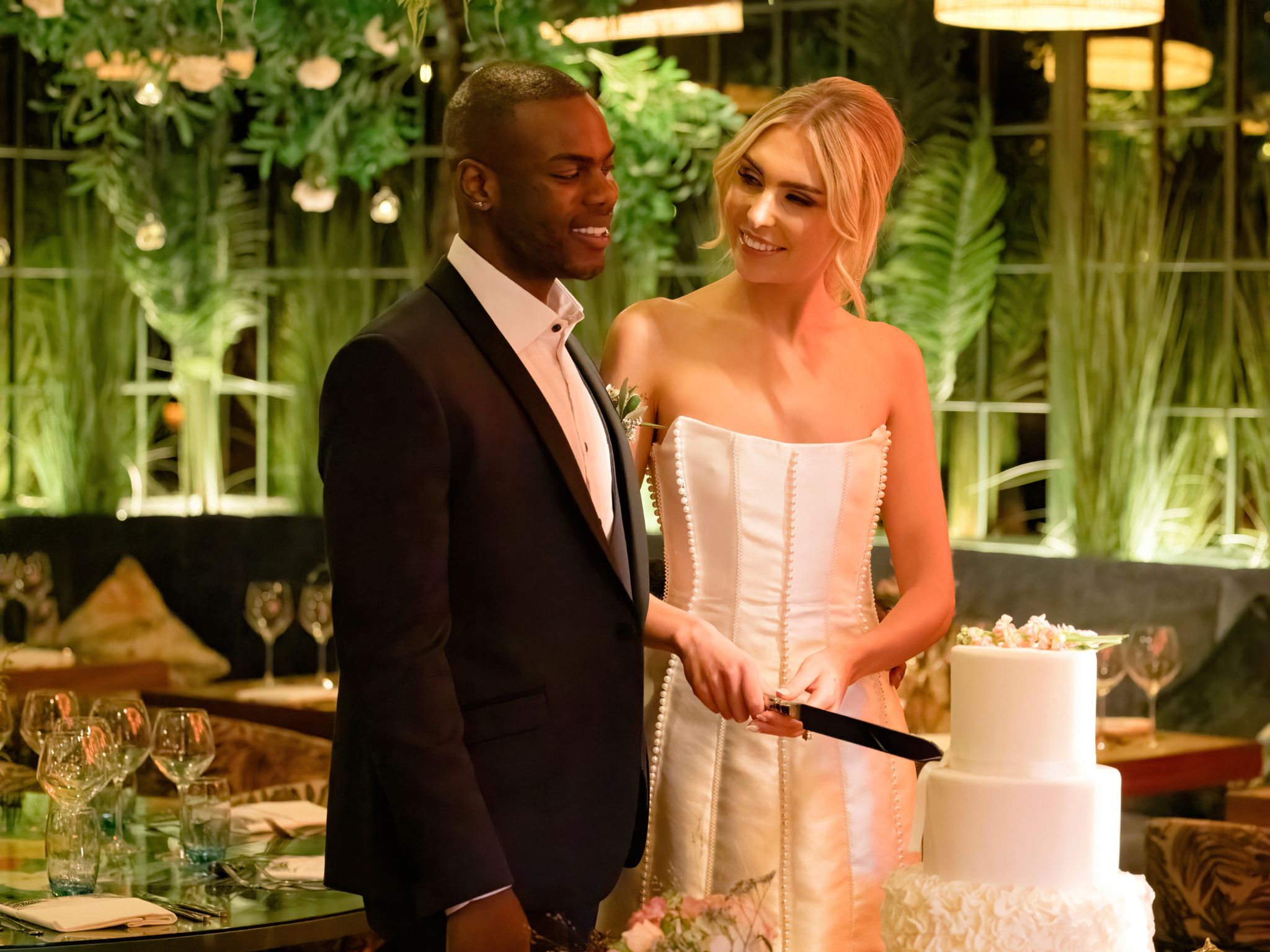 A bride and groom stand together smiling as they cut a white tiered wedding cake in a warmly lit room decorated with lush greenery.