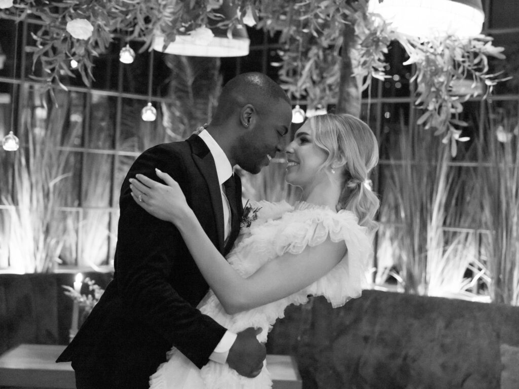 A couple dressed in wedding attire embrace closely and smile at each other while dancing in a softly lit room decorated with hanging greenery and lights.