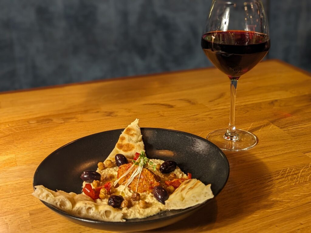 A plate of hummus garnished with olives, roasted peppers, and pita bread slices on a wooden table beside a glass of red wine, creating a cozy dining atmosphere.