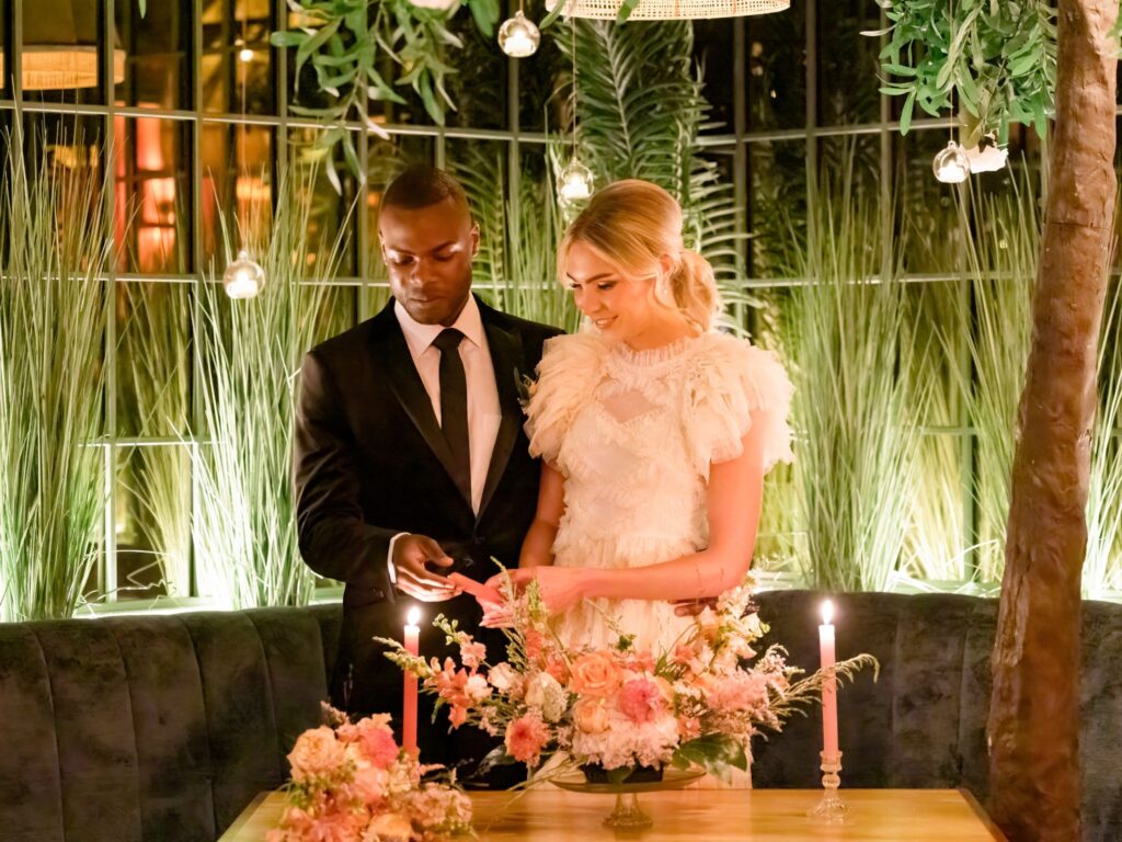 A couple in formal wedding attire stands together at a candlelit table adorned with pink and peach floral arrangements; they appear to be lighting a candle in a warmly lit room filled with tall greenery and hanging lights.