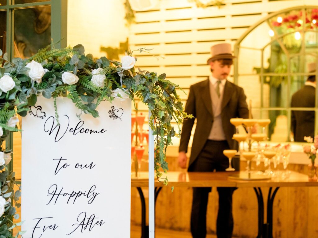 Floral-decorated sign reading ‘Welcome to our Happily Ever After’ displayed at a wedding reception entrance. In the background, a server dressed in a vintage-style suit and top hat stands behind a table arranging champagne glasses on a tiered display, with warm, ambient lighting throughout the venue.