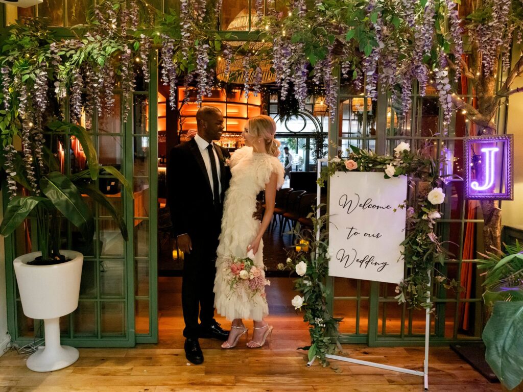 Bride and groom stand together at the entrance of a lush, flower-adorned wedding venue. They face each other, smiling, beneath cascading wisteria and greenery. A large sign beside them reads ‘Welcome to our Wedding,’ decorated with roses and foliage, with warm indoor lighting glowing behind the open doors.