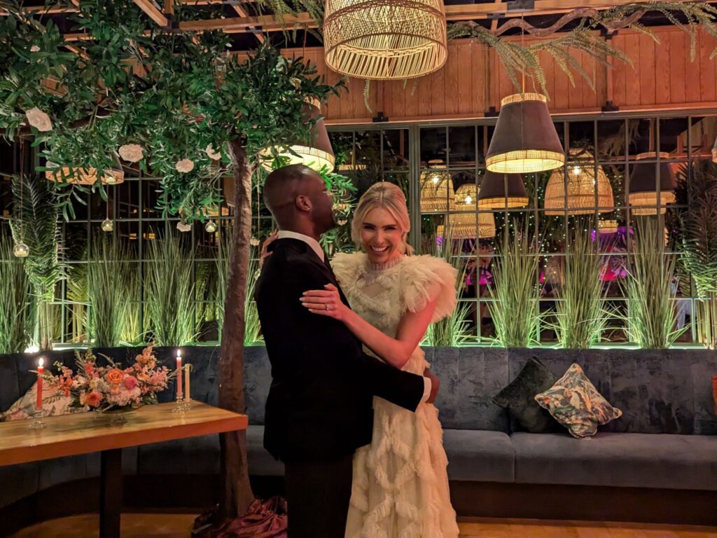 A bride and groom embrace and smile in a warmly lit, lush indoor venue decorated with hanging greenery, woven pendant lights, tall grasses, and candles on a nearby table. The groom wears a dark suit and the bride wears a ruffled ivory gown.