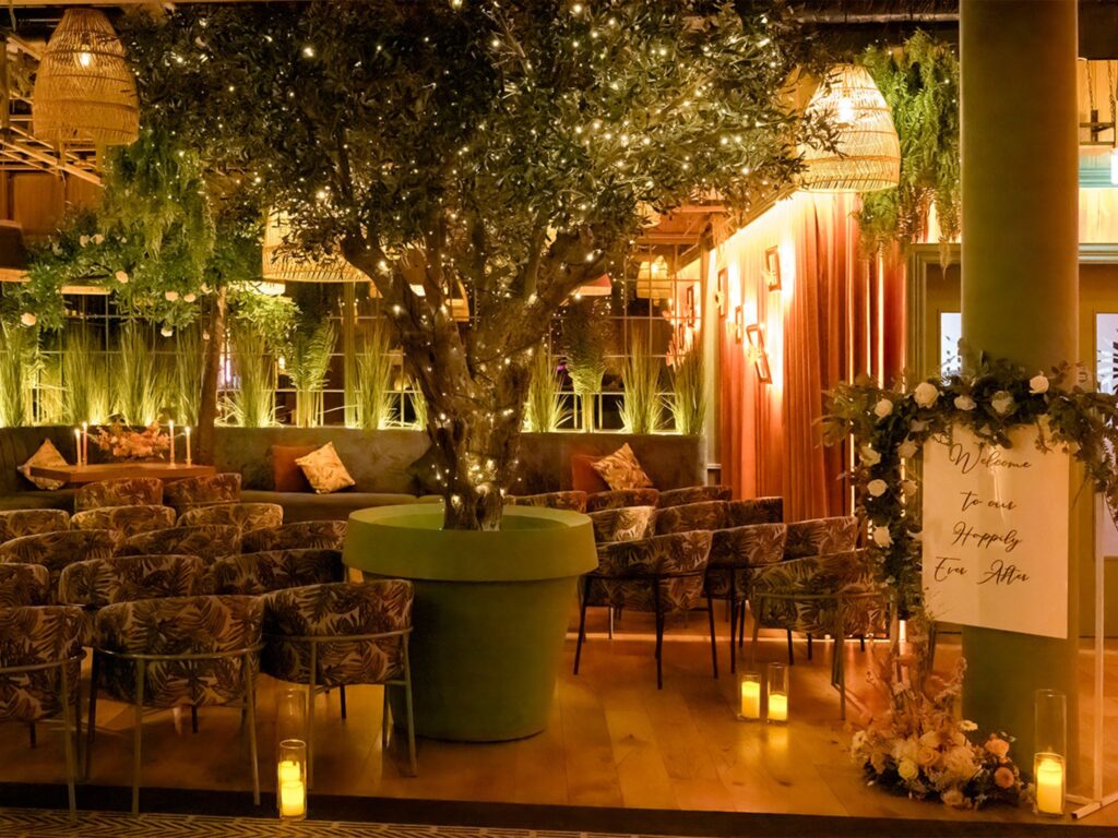 Warmly lit indoor wedding ceremony space featuring rows of botanical-patterned chairs arranged around a large potted tree wrapped in twinkling fairy lights. Lush greenery, soft amber lighting, and hanging woven lamps create a romantic, nature-inspired atmosphere. A decorative sign at the entrance reads ‘Welcome to our Happily Ever After,’ surrounded by floral arrangements and candles.