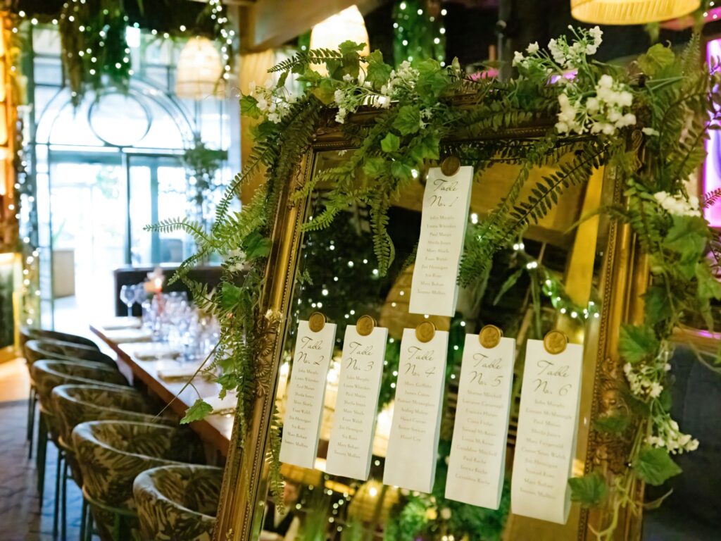 An ornate gold-framed mirror decorated with greenery and white flowers displays wedding table assignment cards, each clipped with a gold seal. Behind it, a warmly lit reception area features a long dining table set with glassware, surrounded by chairs with botanical-patterned upholstery and lush greenery throughout the space.