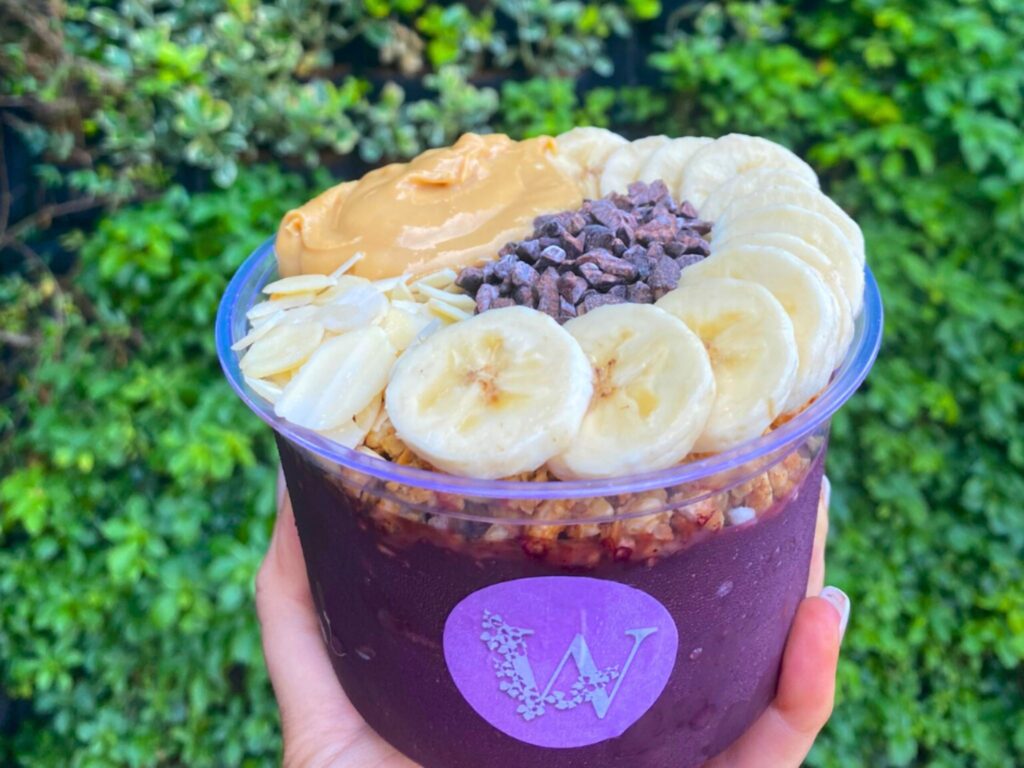 A hand holds a purple acai bowl topped with sliced bananas arranged in a ring, almond flakes, cacao nibs, granola, and a large spoonful of peanut butter. The bowl has a purple label with a floral-style “W” logo, and the background is filled with lush green foliage.