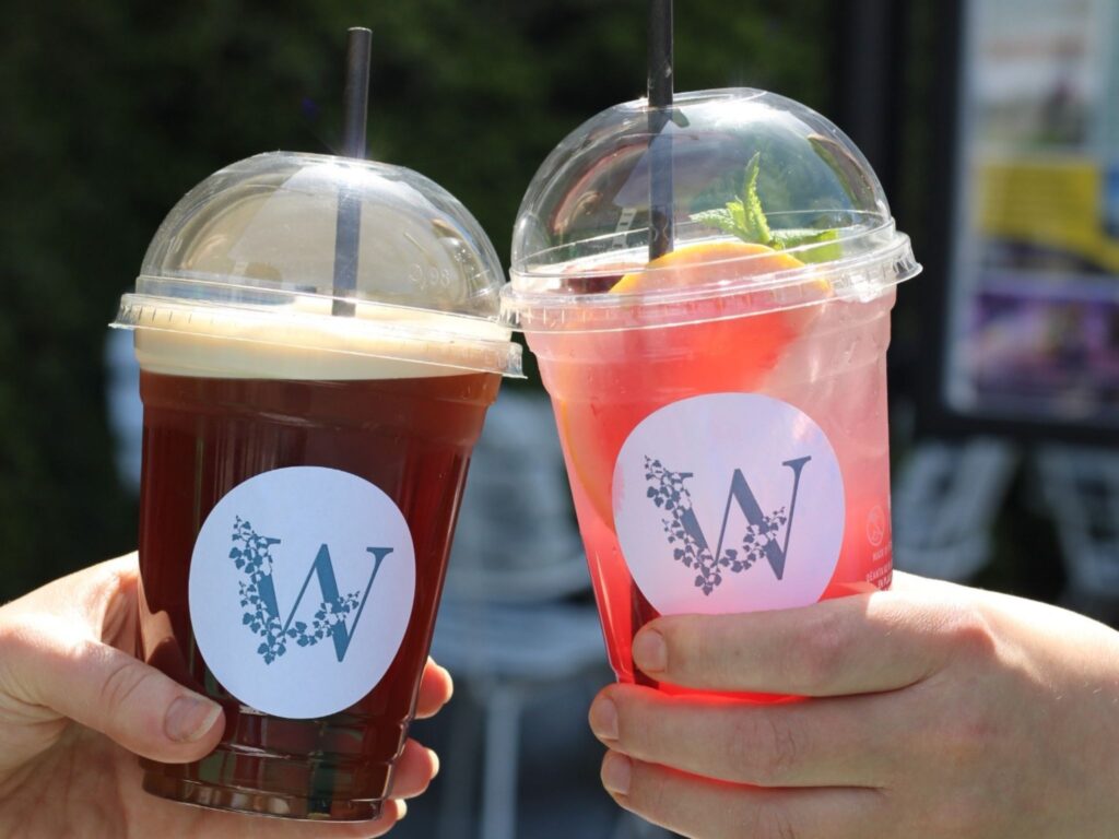 Two hands hold up iced drinks in clear plastic cups with dome lids and black straws. The left drink is a dark iced coffee or tea, and the right drink is a bright pink lemonade with lemon slices and mint. Both cups display a white sticker with a stylized “W” logo made of small floral elements. The background is outdoors and slightly blurred.