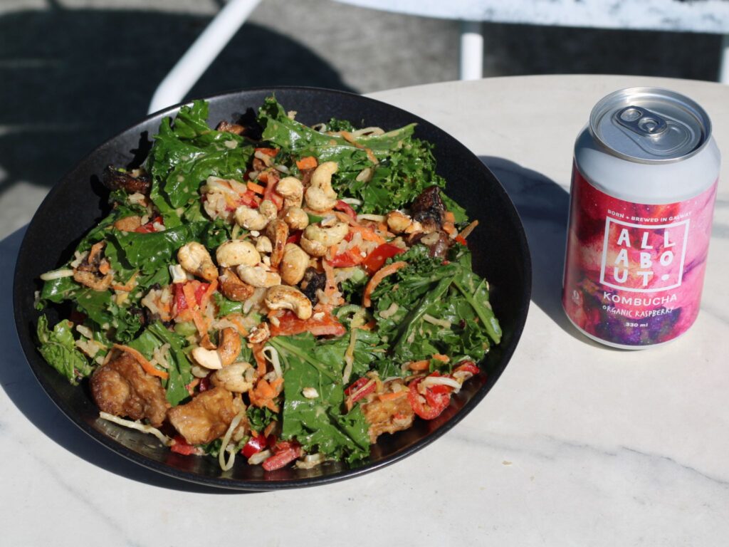 A black bowl filled with a colorful salad containing kale, vegetables, cashews, and pieces of cooked chicken or tofu sits on a white outdoor table. Next to the bowl is a can of All About Kombucha in an organic raspberry flavor, with a pink and purple label.