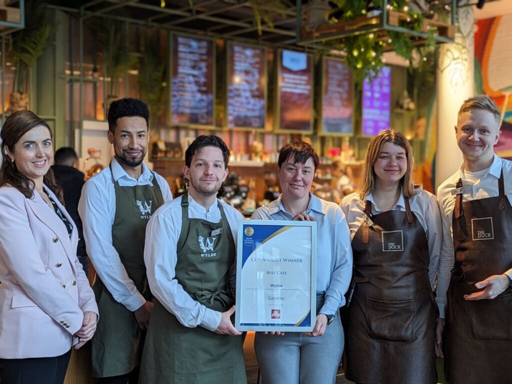 A group of six café staff members stand together indoors, smiling and posing for a photo. The person in the center holds a framed award certificate that reads “Connaught Winner – Best Café – Wylde – Galway.” The staff members wear uniforms and aprons, and the background shows a modern café interior with menu screens and plants.