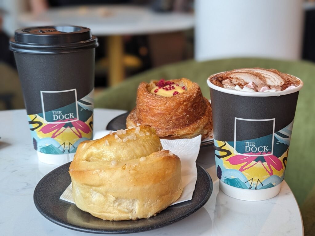 Two takeaway cups of coffee and hot chocolate from “The Dock” sit on a café table alongside two pastries: a glazed swirl bun and a flaky pastry filled with custard and topped with dried raspberries.