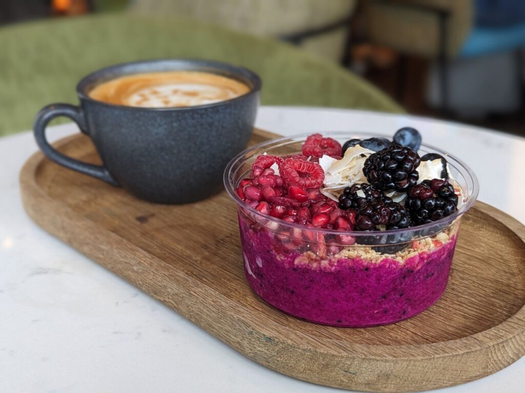A wooden serving tray holding a dark ceramic cup of latte and a vibrant pink smoothie bowl topped with granola, pomegranate seeds, raspberries, blackberries, blueberries, and coconut flakes, placed on a white table with soft seating in the background.