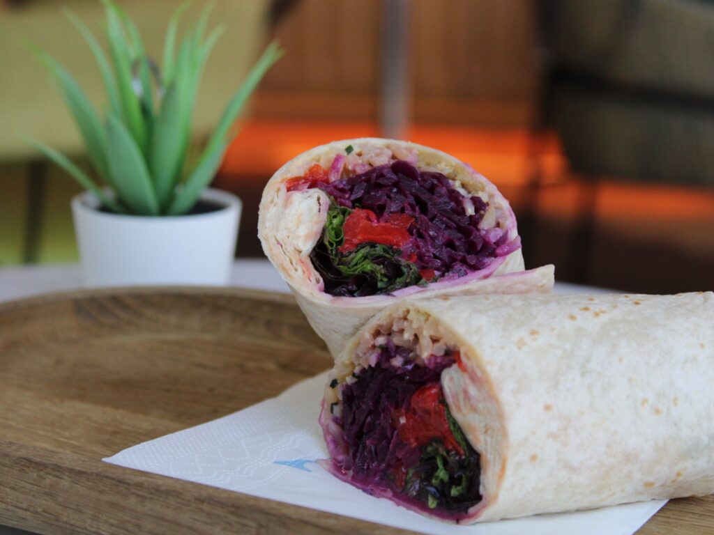 A wrap cut in half and stacked, filled with brightly colored vegetables including red cabbage, roasted red peppers, and leafy greens, placed on a wooden tray with a small potted plant in the background.