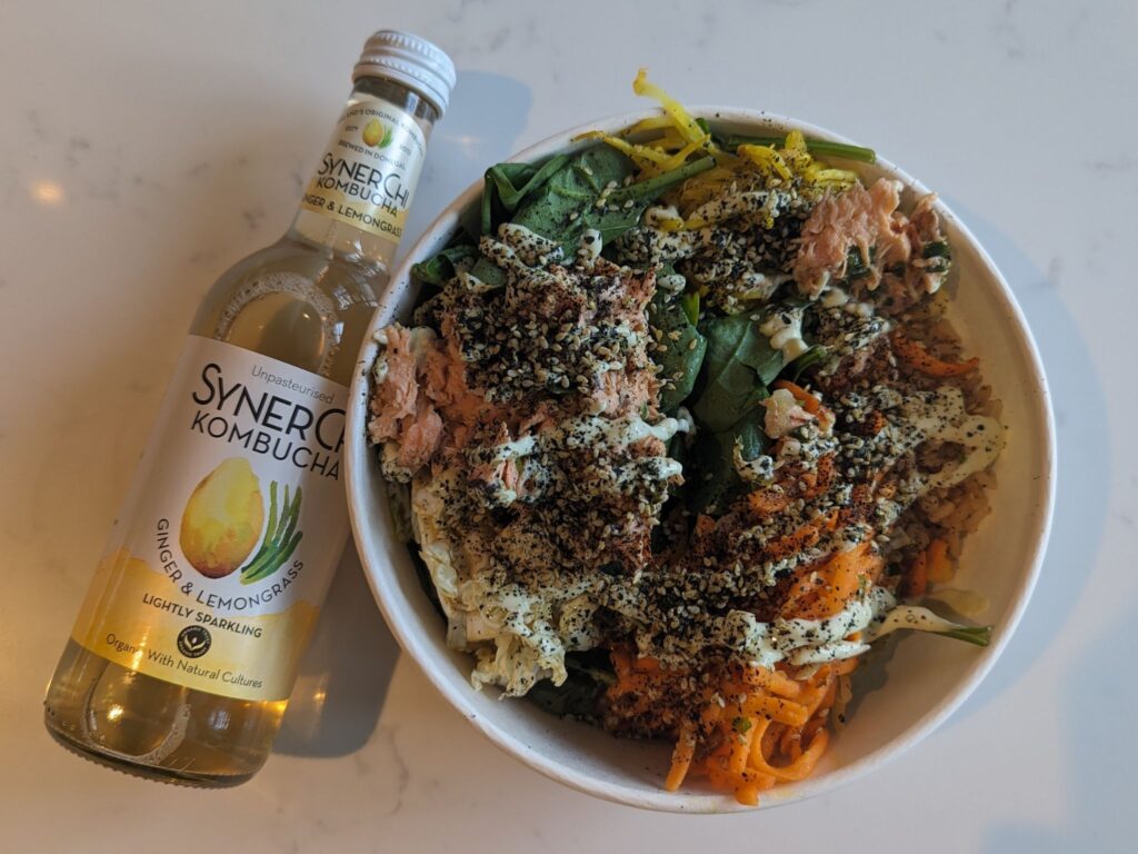 A bowl filled with a mixed salad containing shredded vegetables, spinach, grains, and flaked salmon topped with herbs and dressing, placed beside a bottle of SynerChi ginger and lemongrass kombucha on a white surface.
