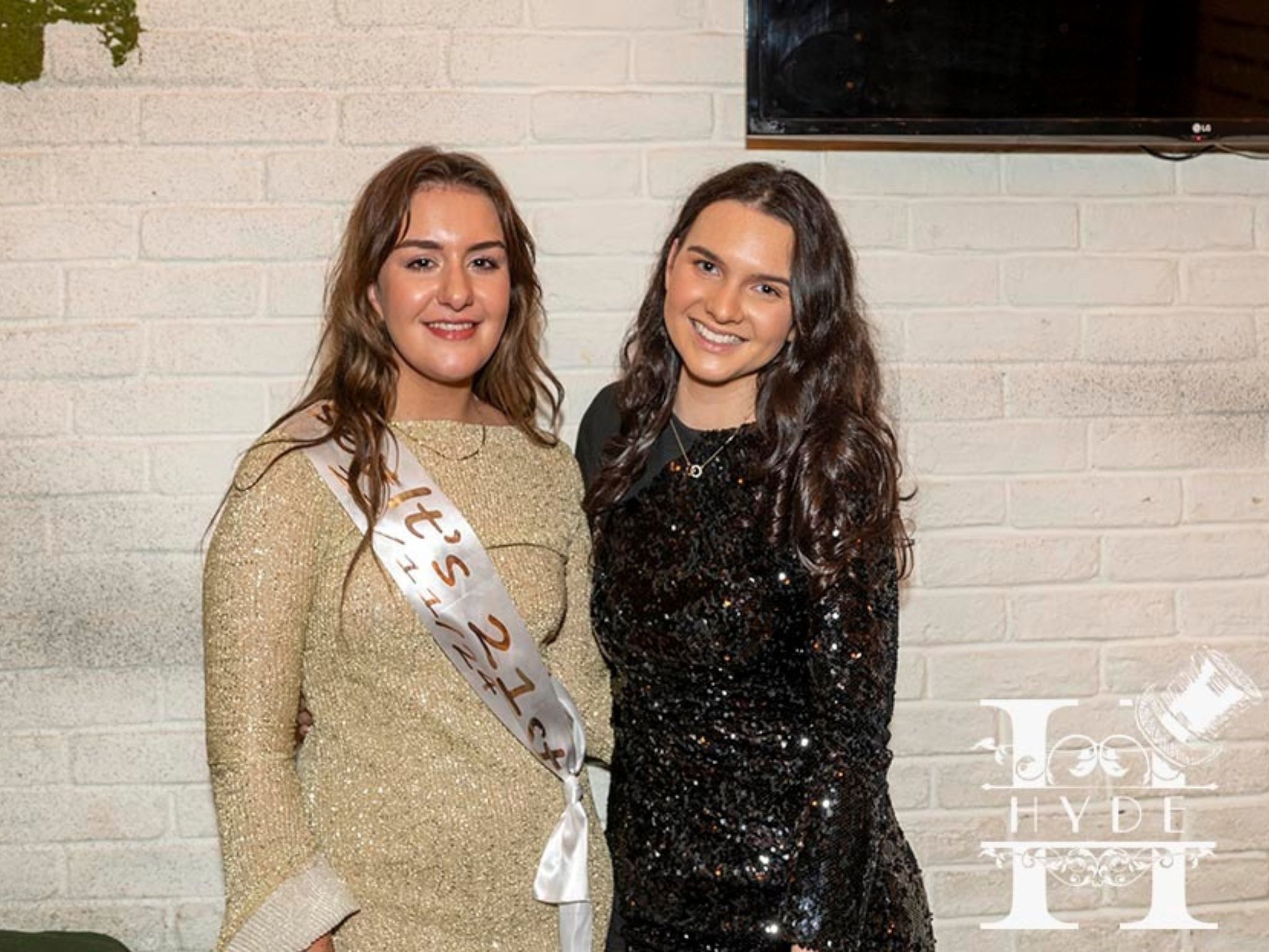 Two women smiling in front of a white brick wall. One wears a glittery gold dress with a "21st" sash, the other wears a black sequin dress. Festive and joyful atmosphere.