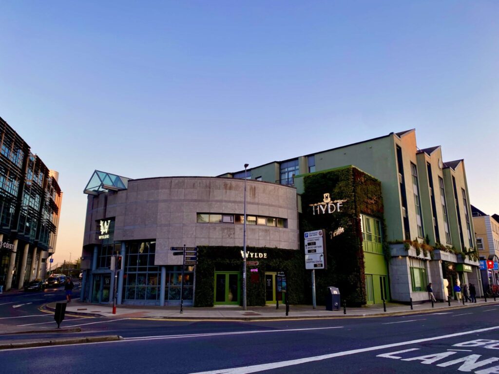 A street-corner view of a modern building complex at sunset, featuring the WYLDE café with green ivy-covered walls and large windows, adjacent to the HYDE hotel building. The sky is clear and the street in the foreground shows road markings, pedestrians, and nearby city buildings.