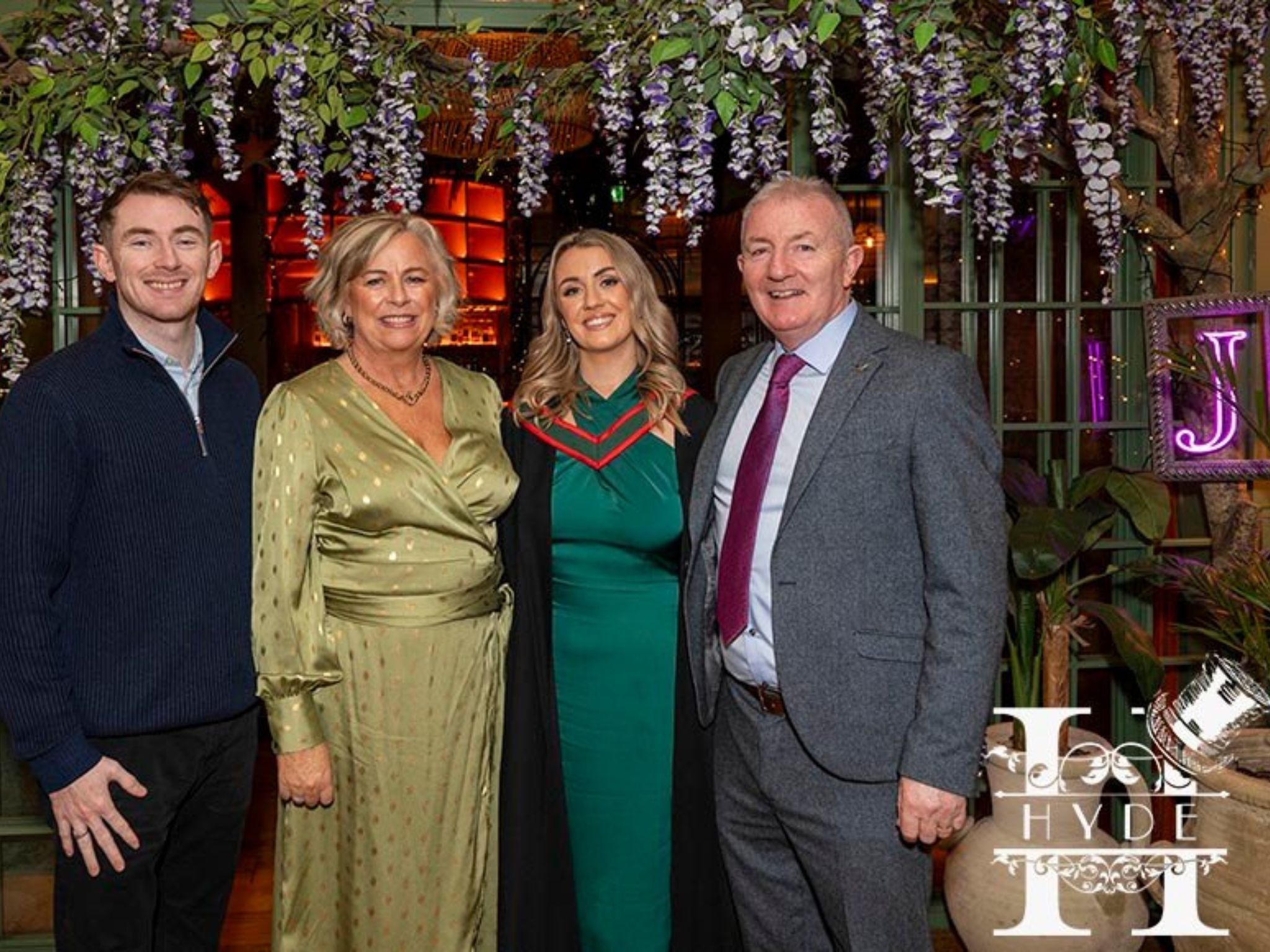 A joyful family portrait at an event, featuring two men and two women smiling. The women wear a green dress and a graduation gown. Lush greenery and purple flowers adorn the background, with warm lighting adding a celebratory atmosphere.